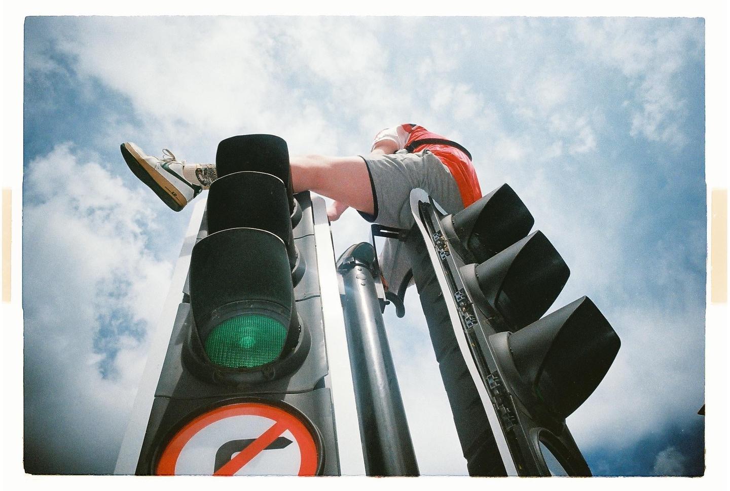 A person is sitting on top of a traffic light against a cloudy sky.