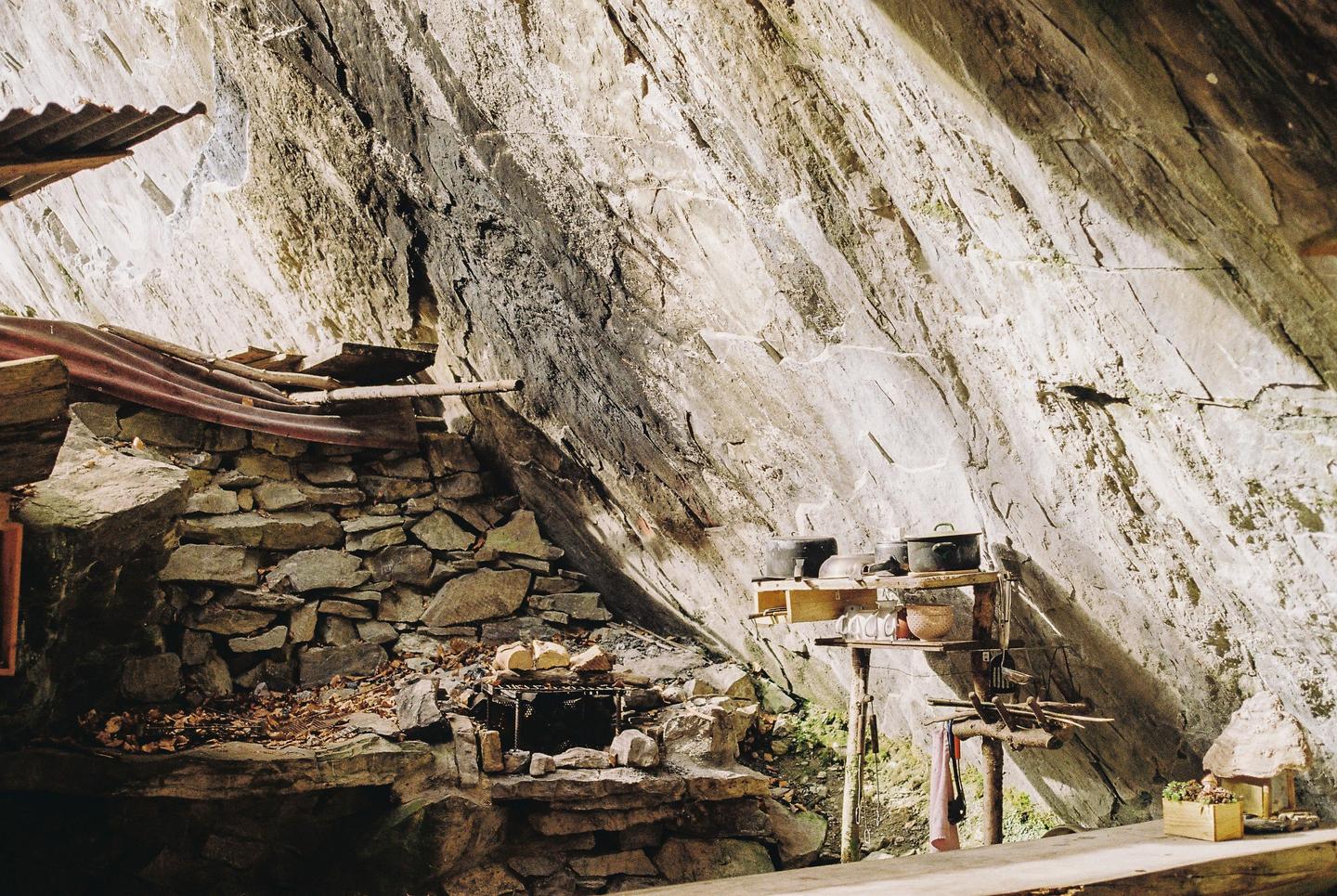 A rustic outdoor kitchen with a stone fireplace and wooden shelves is built into the side of a large rock formation.