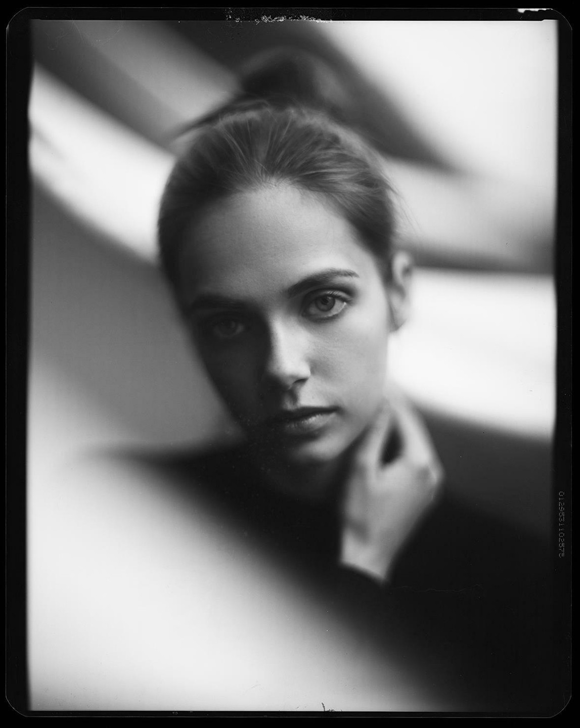 A woman with her hair in a bun looks directly at the viewer, with one hand touching her neck.