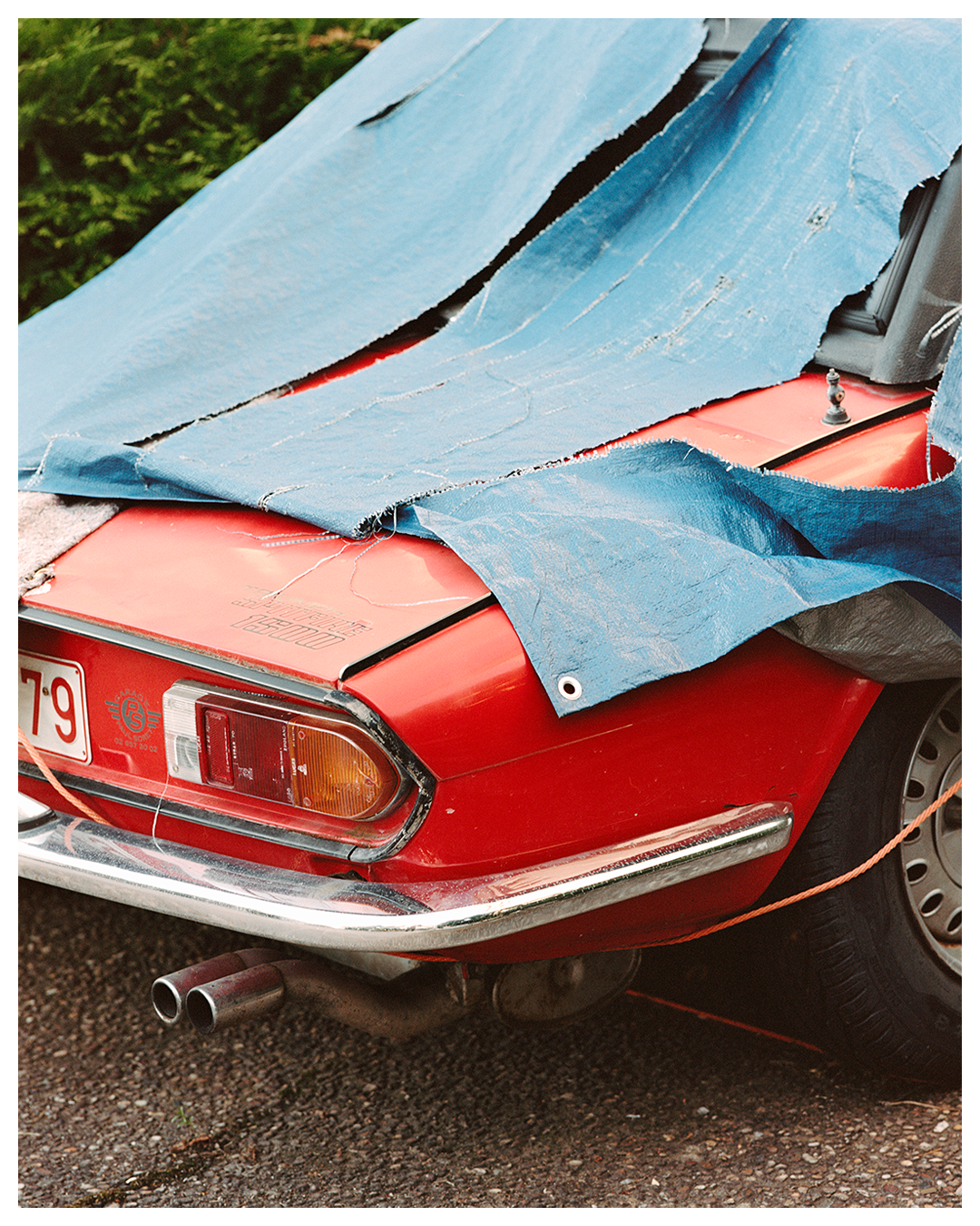 A red car is partially covered by a torn blue tarp.