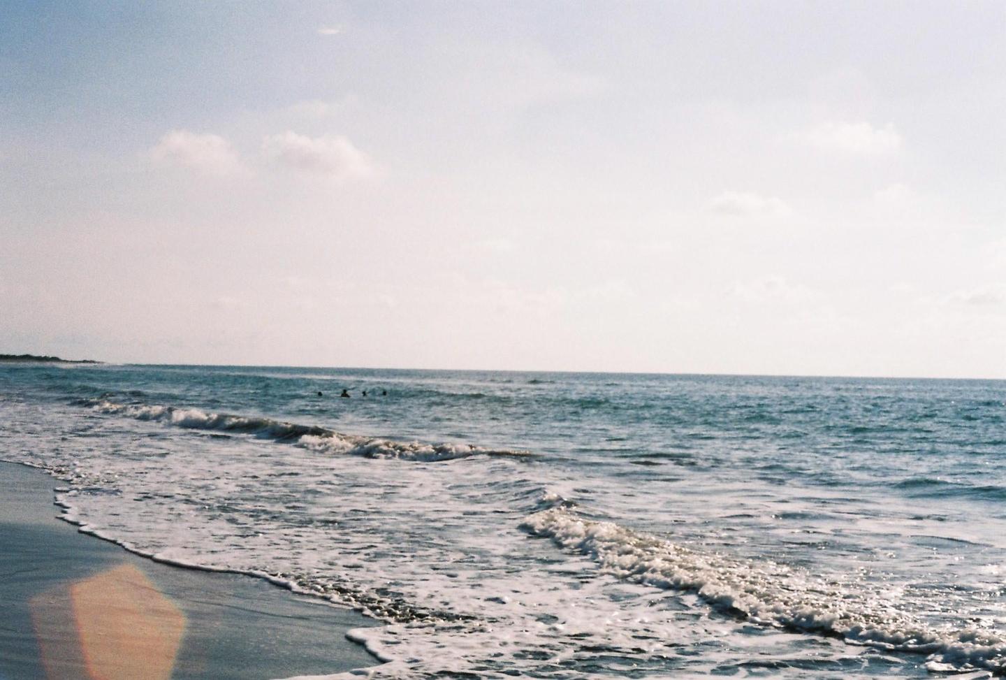 Waves crash on a sandy beach with a few people swimming in the distance under a bright sky.