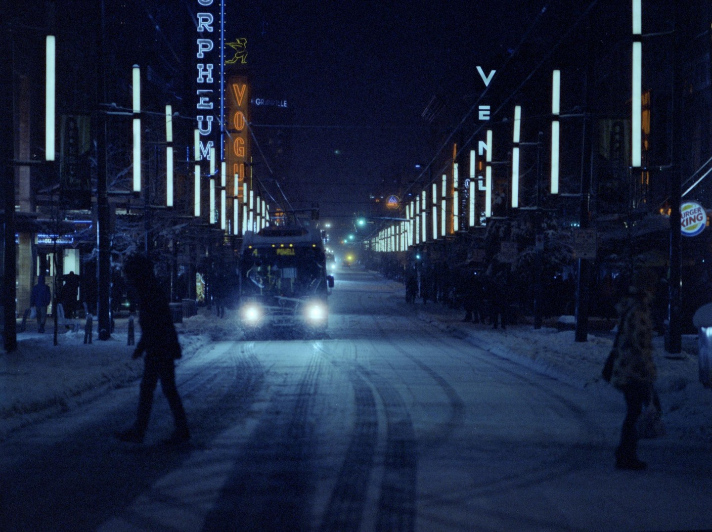 A snow-covered street at night is illuminated by neon signs and a bus with bright headlights.