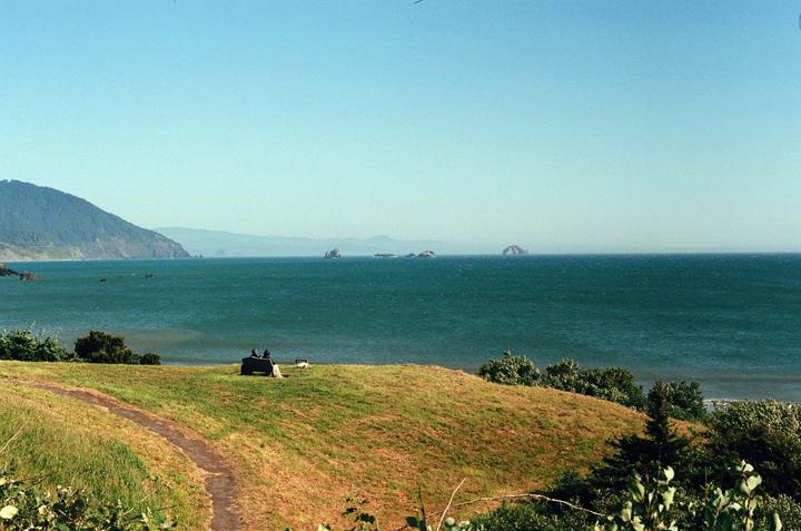A couple sits on a bench overlooking the ocean with mountains and sea stacks in the distance.