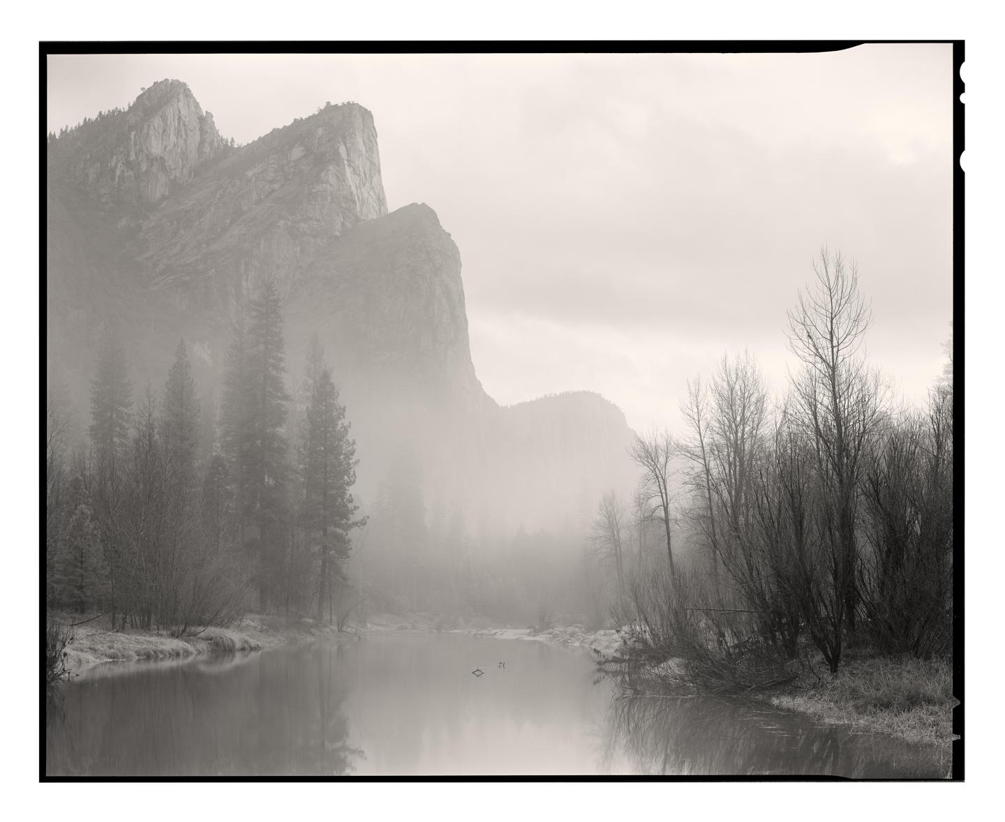 A misty mountain landscape features a river reflecting the sky and bare trees on its banks.