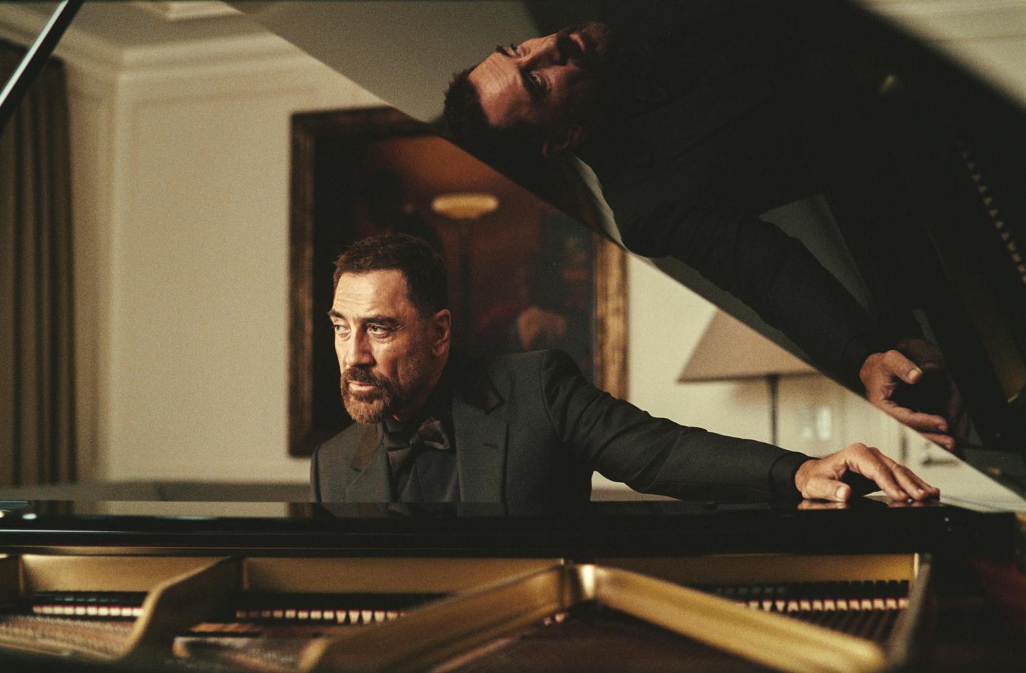 A man in a suit sits at a grand piano, with his reflection visible in the piano's lid.