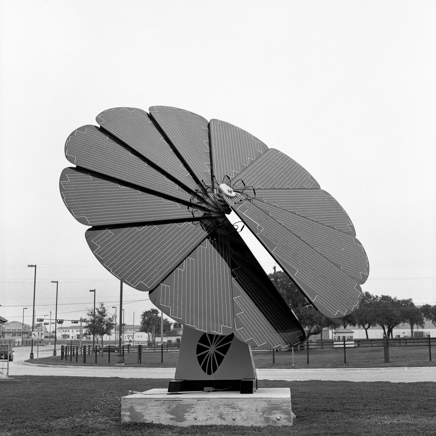 A large, flower-shaped solar panel stands in a grassy area next to a road.