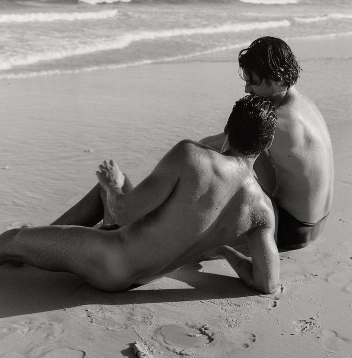 Two men are sitting on a sandy beach next to the ocean.