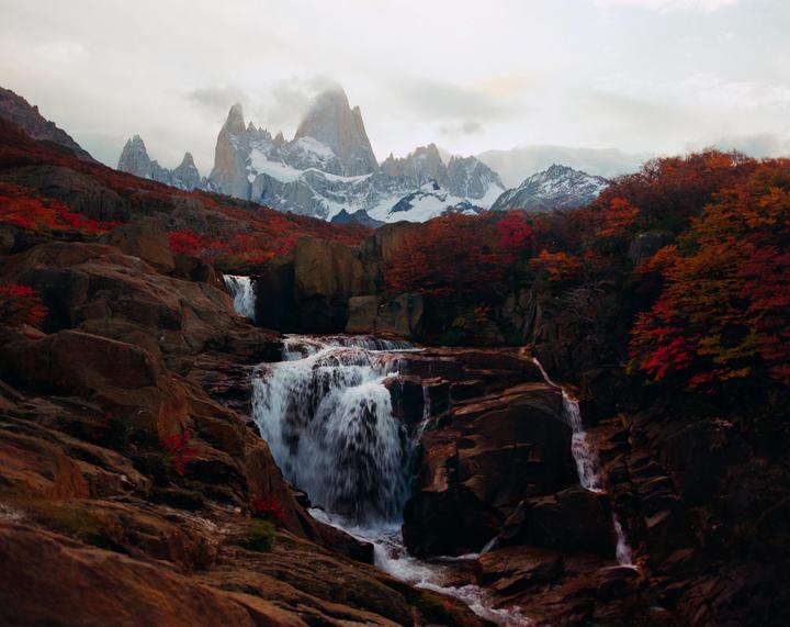 A waterfall cascades over rocks with snow-capped mountains and autumn foliage in the background.