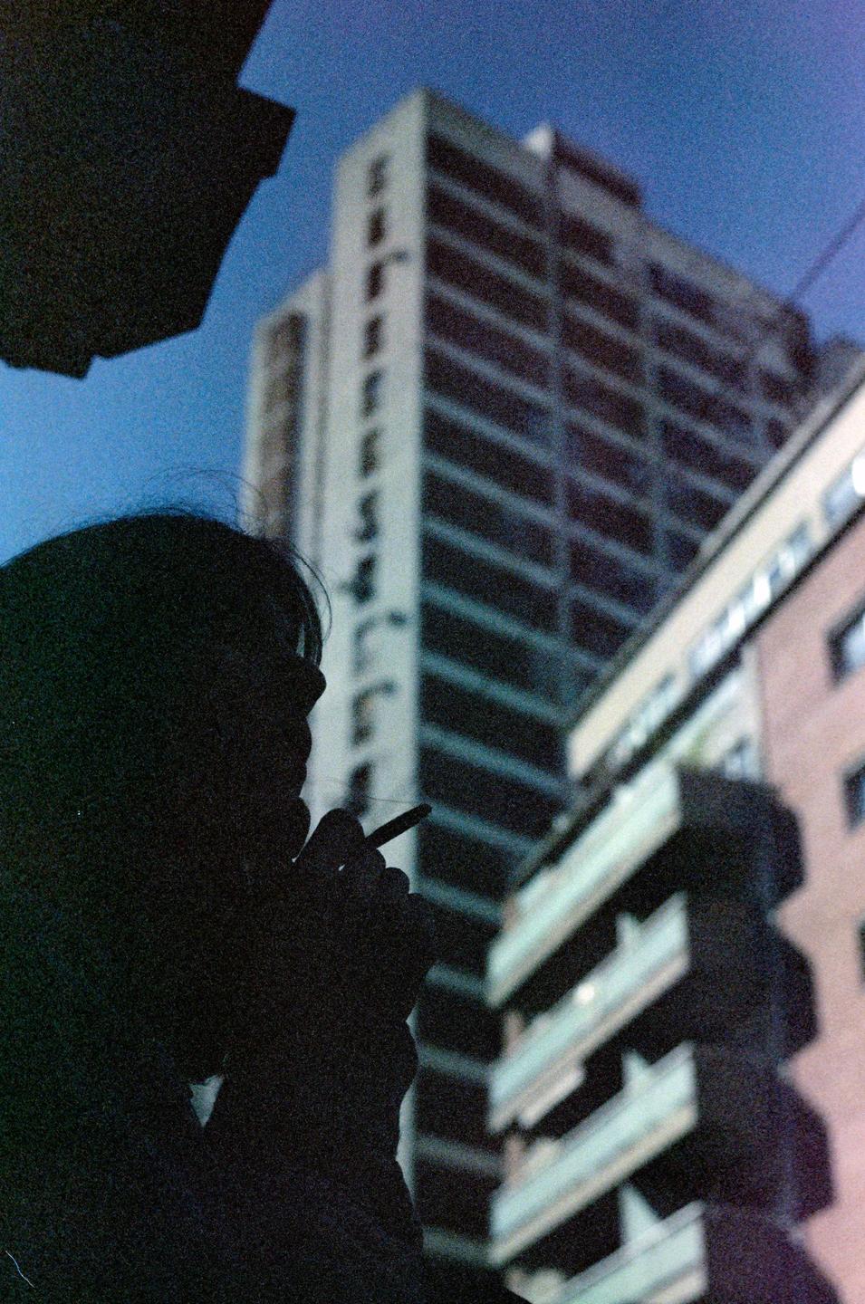 A silhouette of a woman smoking is visible against a backdrop of tall buildings and a blue sky.