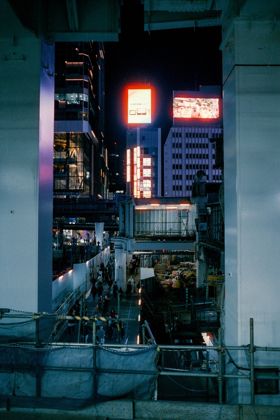 A busy street at night is framed by two large pillars, with illuminated buildings and billboards in the background.