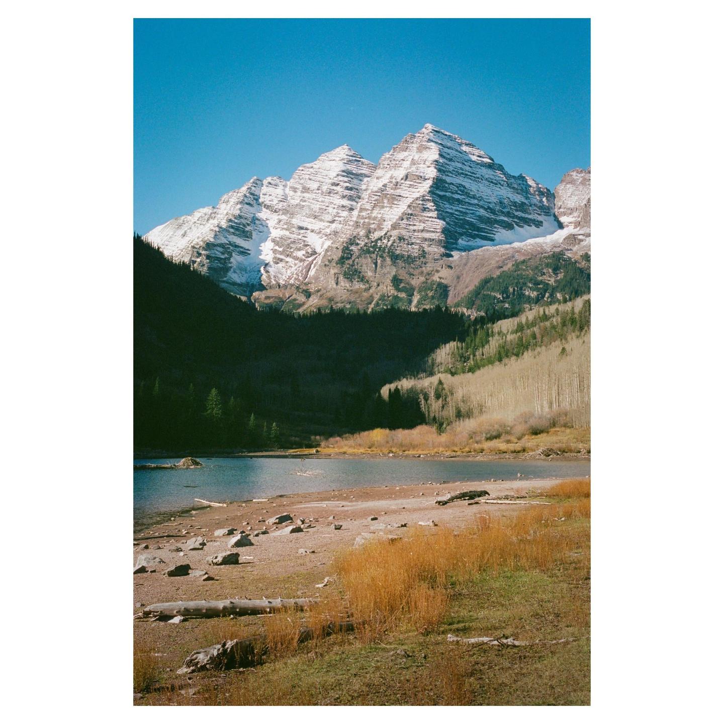A lake with a rocky shore is in the foreground, with a forest and snow-capped mountains in the background.
