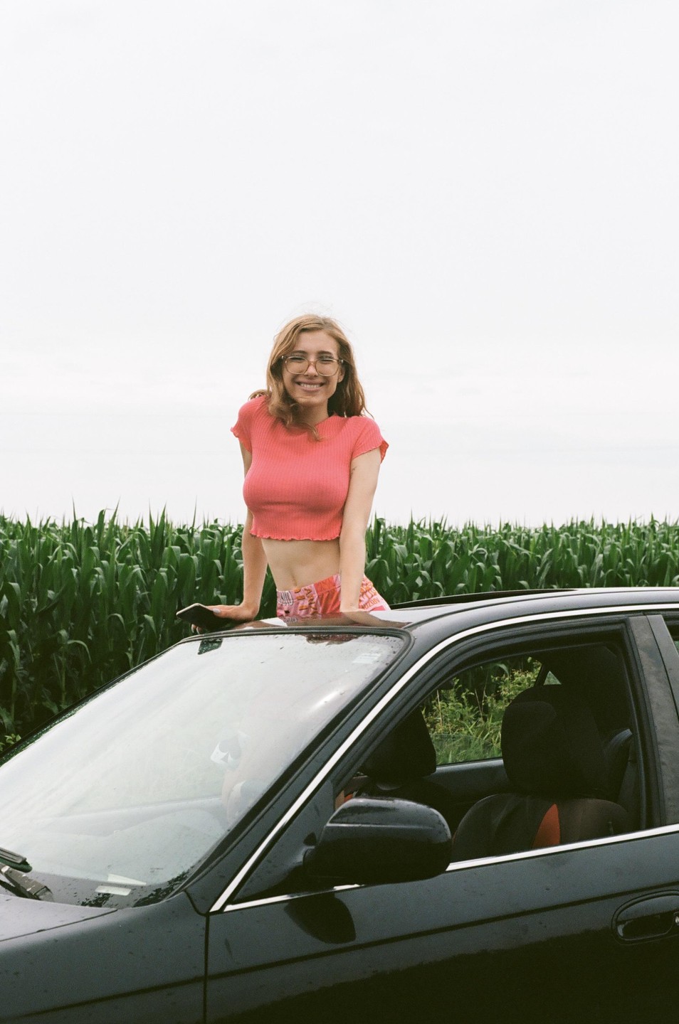 A smiling woman with glasses leans out of a car sunroof next to a field of tall green crops.