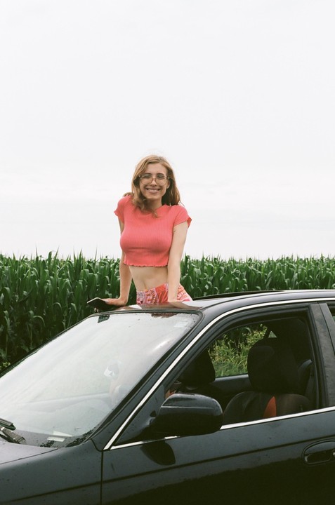 A smiling woman with glasses leans out of a car sunroof next to a field of tall green crops.