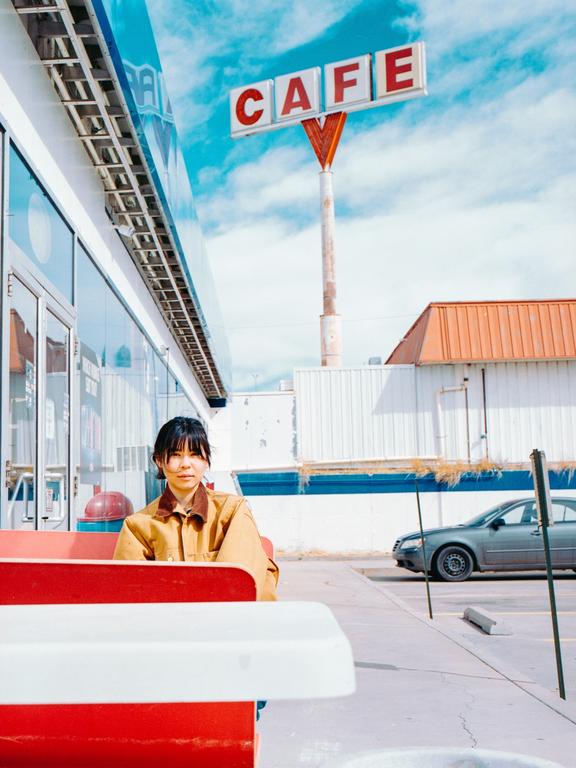 A woman sits at an outdoor cafe table with a large cafe sign and a car in the background.
