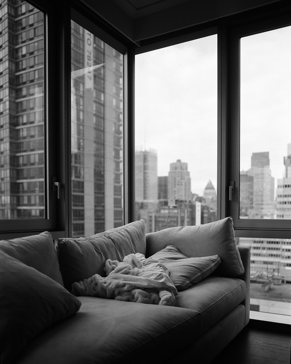 A couch with pillows and a blanket sits by a large window overlooking a city skyline.