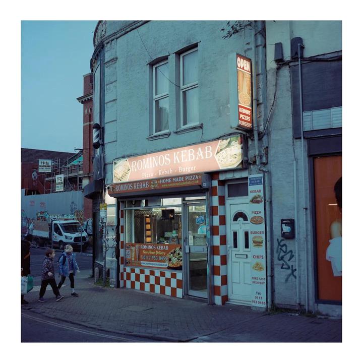 Two children walk past a kebab shop with a checkered facade and a barber pole on the corner of a street.