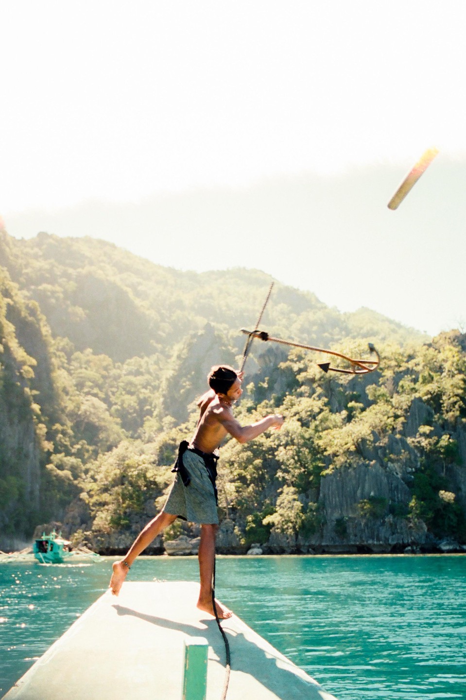 A man stands on the bow of a boat and throws an anchor into the turquoise water with mountains in the background.