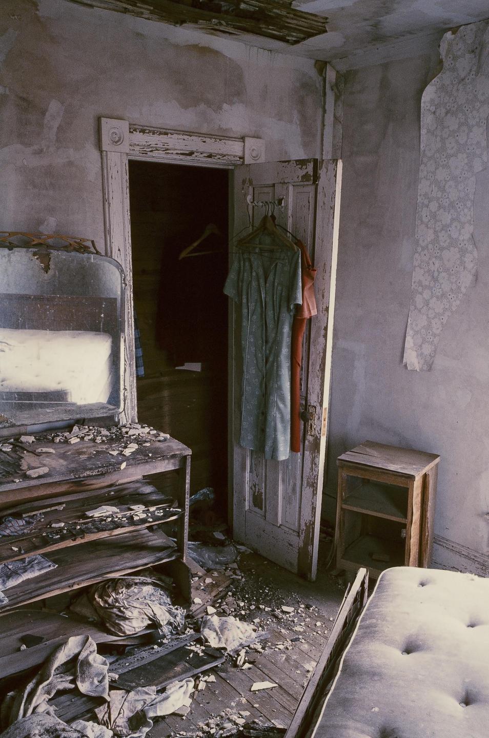 An abandoned room with peeling paint and debris on the floor contains a dresser with a mirror, a bed frame, and a door with clothes hanging on it.