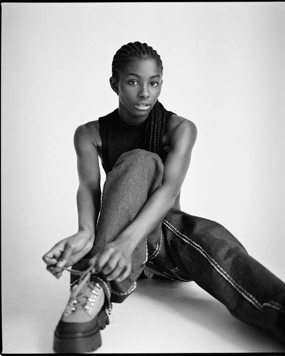 A woman with braided hair sits on the floor and ties the laces of her shoe.