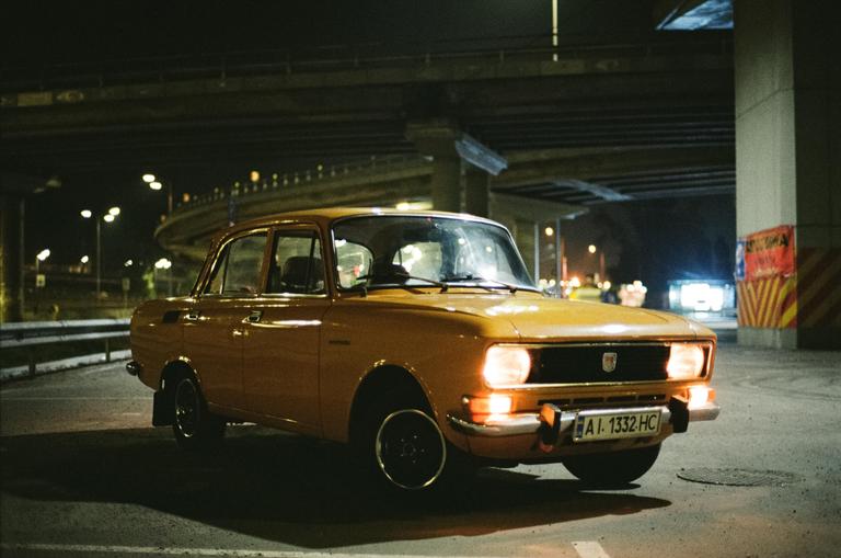 A yellow car with its headlights on is parked under an overpass at night.