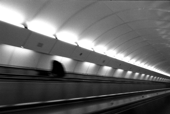 A blurry figure stands on an escalator in a brightly lit tunnel.