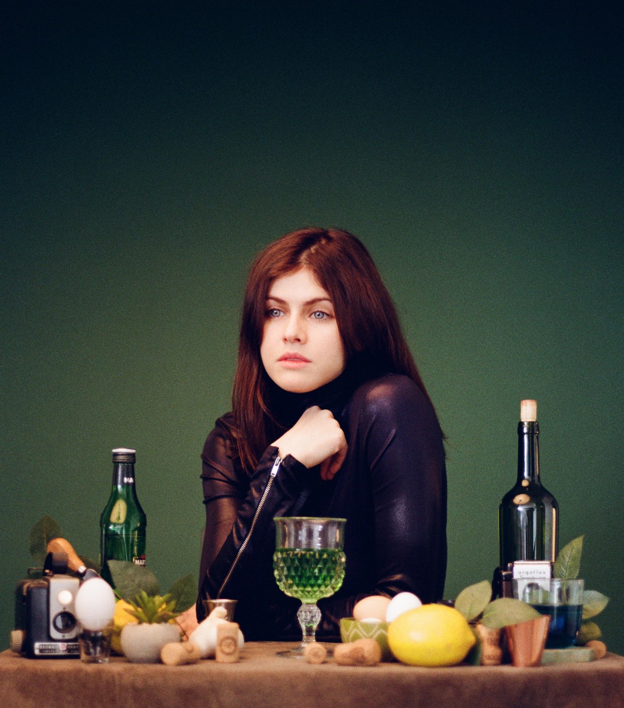 A woman with long brown hair and blue eyes sits at a table with bottles, glasses, and fruit.