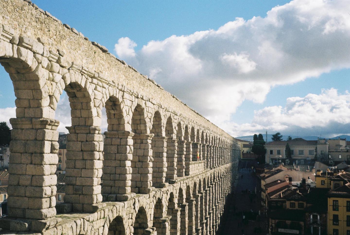 A long stone aqueduct with many arches stretches across the frame under a blue sky with white clouds, overlooking a town.