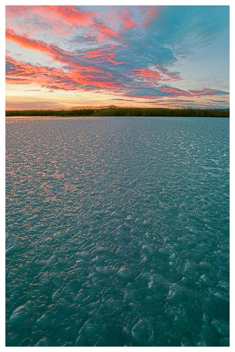 A frozen lake with a line of reeds in the distance under a colorful sunset sky.