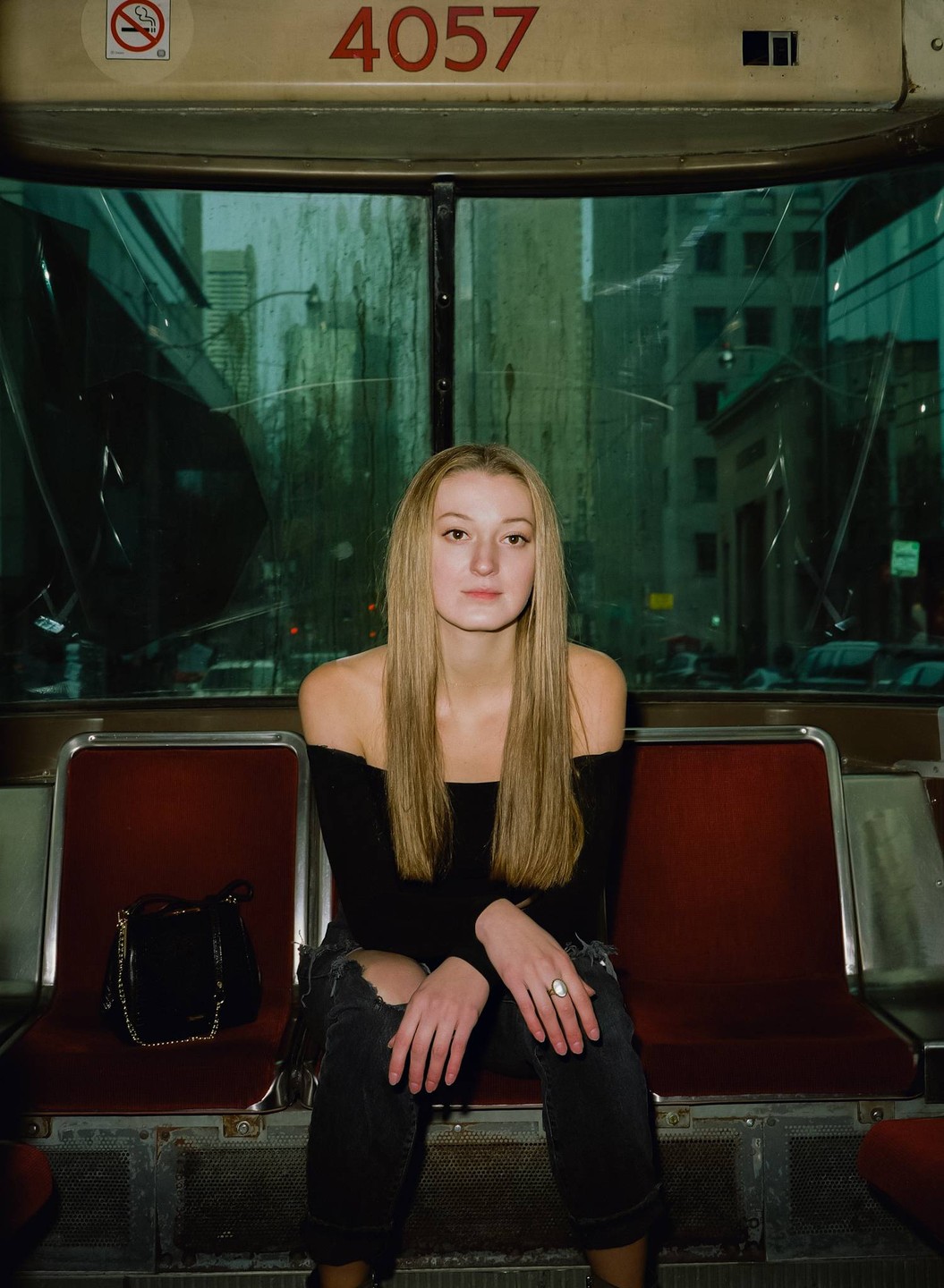 A woman with long blonde hair sits on a red seat inside a vehicle, looking directly at the viewer.
