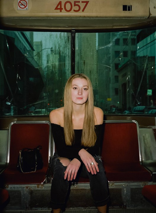 A woman with long blonde hair sits on a red seat inside a vehicle, looking directly at the viewer.