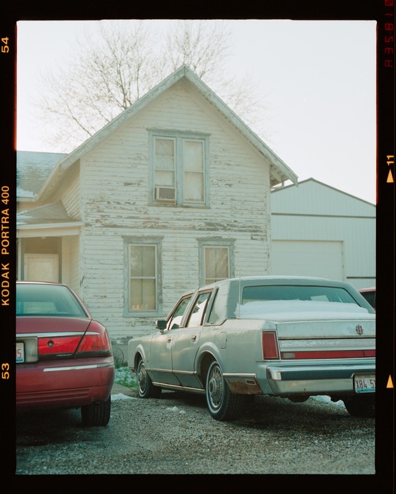 Two cars are parked on a gravel driveway in front of a weathered house with peeling paint.