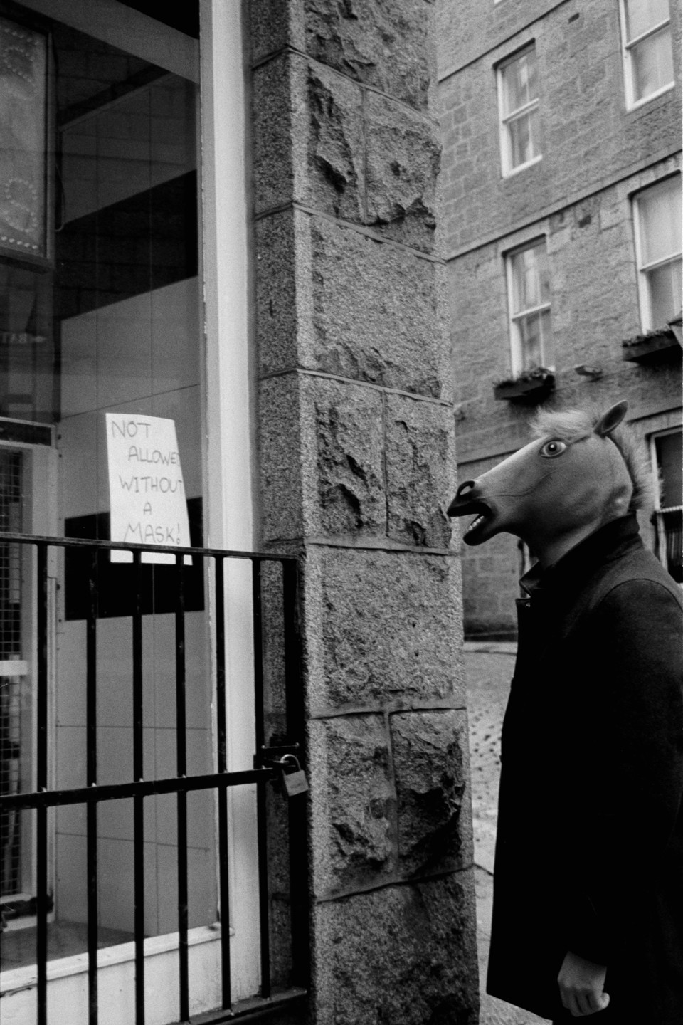 A person wearing a horse mask stands next to a building with a sign that reads "NOT ALLOWED WITHOUT A MASK!".