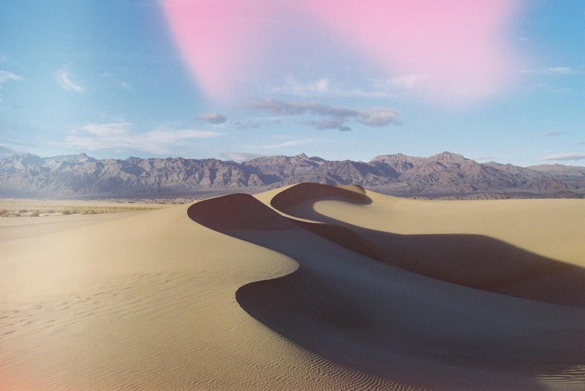 A desert landscape shows sand dunes with long shadows and mountains in the distance under a blue sky with a pink light leak.
