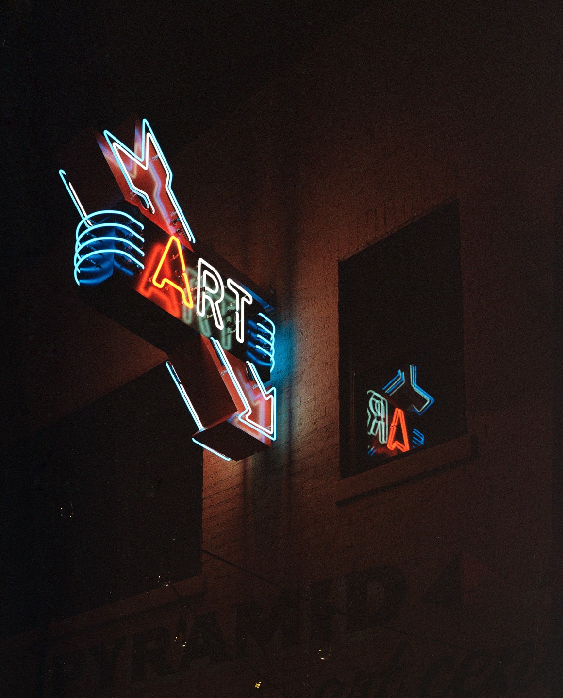 A neon sign with the word "ART" and an arrow points down, with its reflection visible in a window.