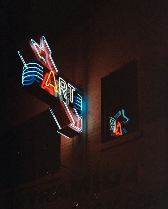 A neon sign with the word "ART" and an arrow points down, with its reflection visible in a window.