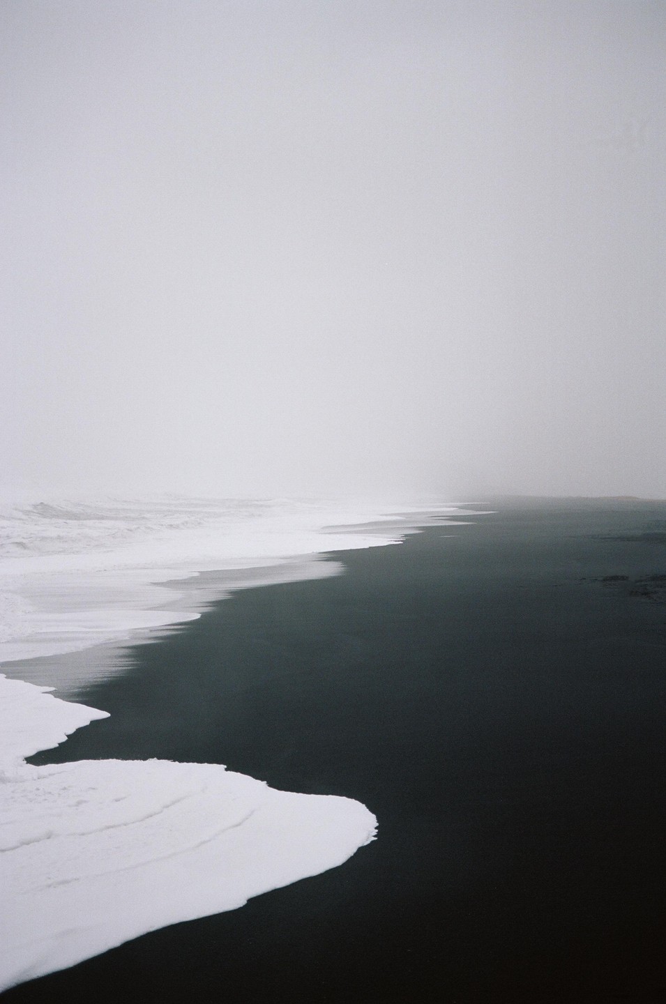 Waves crash on a black sand beach under a hazy sky.