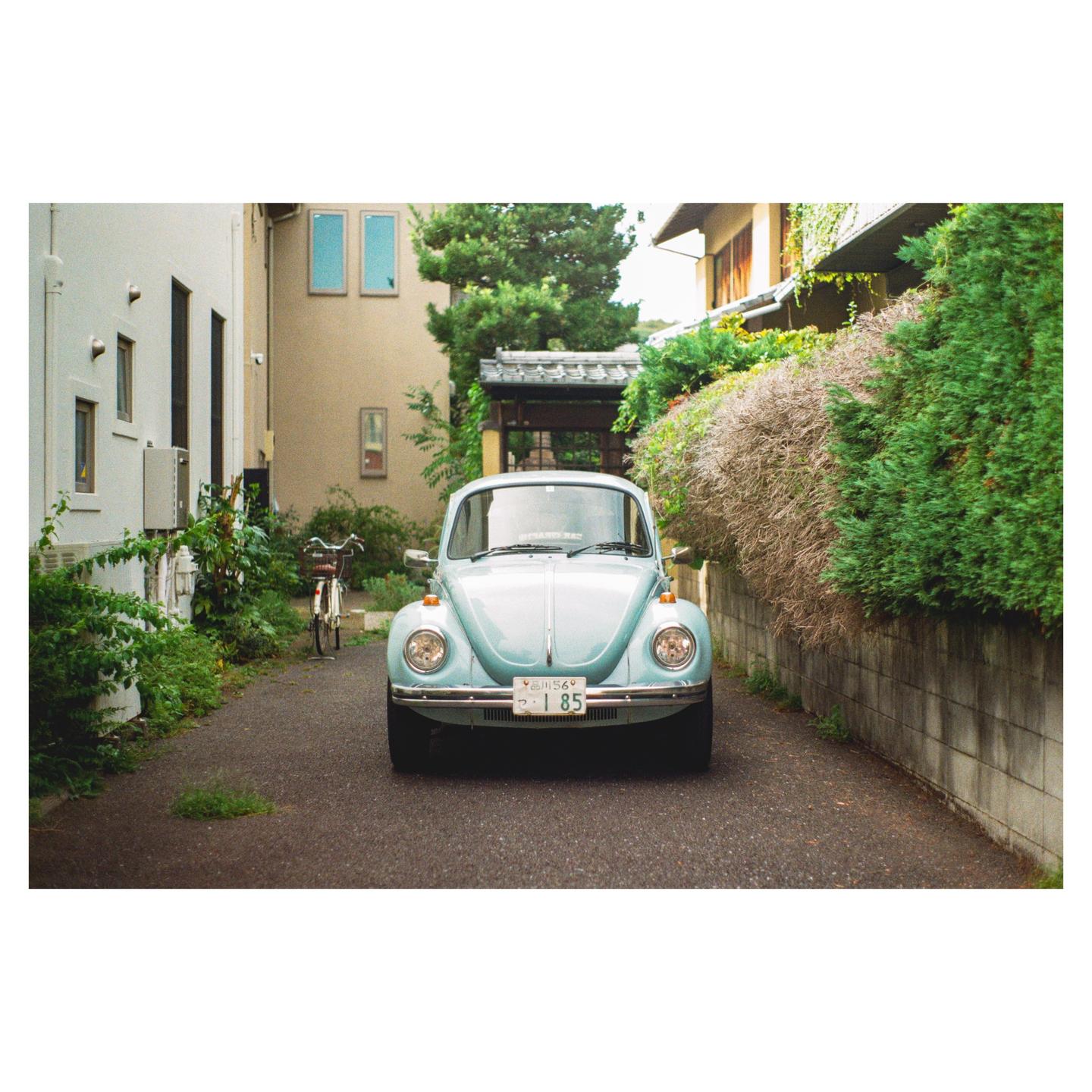 A light blue vintage car is parked on a narrow paved road between buildings and lush green foliage.