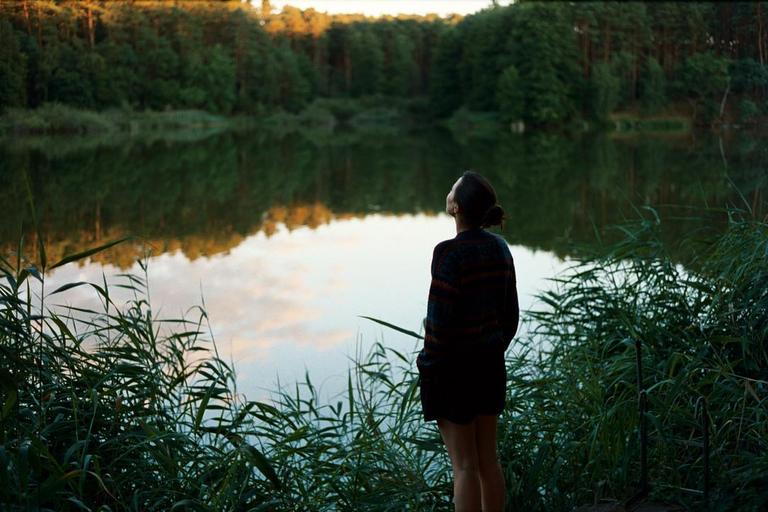 A woman stands by a lake surrounded by tall grass and trees, looking up at the sky.