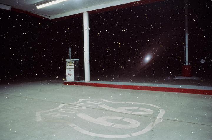 A gas station with a Route 66 logo on the ground is shown under a starry night sky with a visible galaxy.