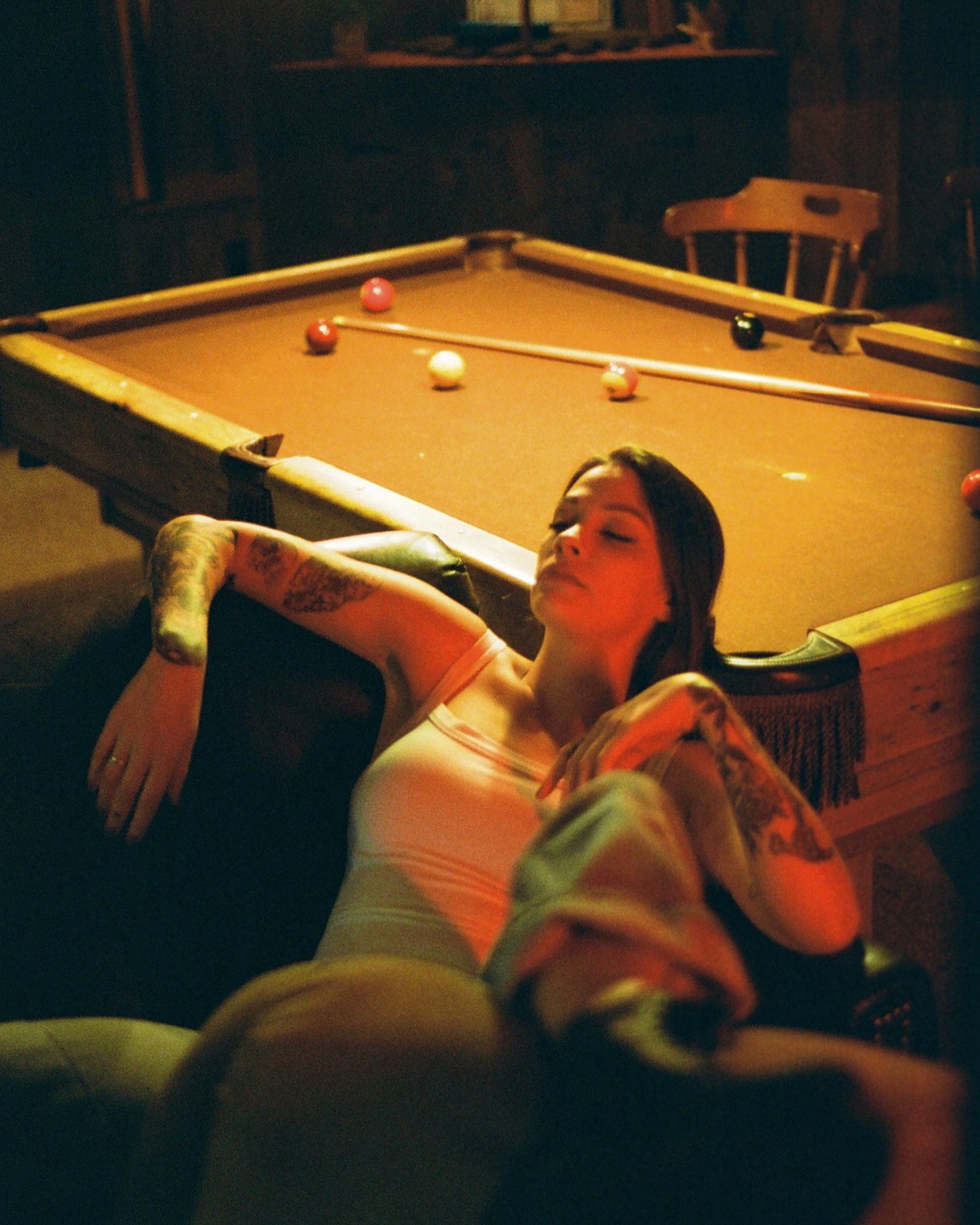 A woman with tattoos on her arms is relaxing on a couch next to a pool table.