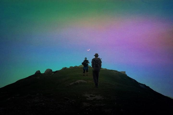 Two people with backpacks walk up a grassy hill under a colorful sky with a crescent moon.