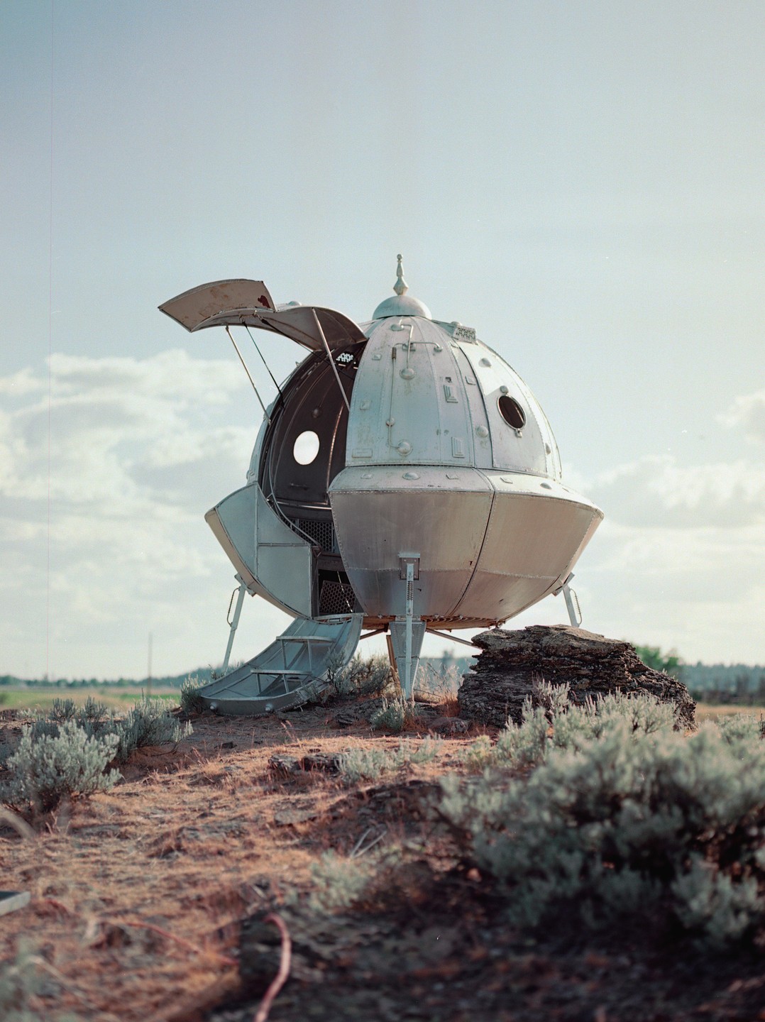 A silver, spherical spaceship with an open hatch and deployed ramp sits on a dry, scrubby hill under a partly cloudy sky.