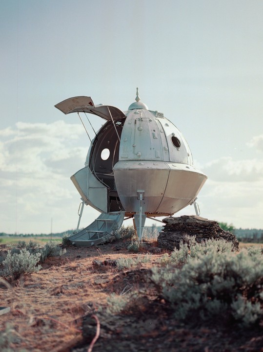 A silver, spherical spaceship with an open hatch and deployed ramp sits on a dry, scrubby hill under a partly cloudy sky.