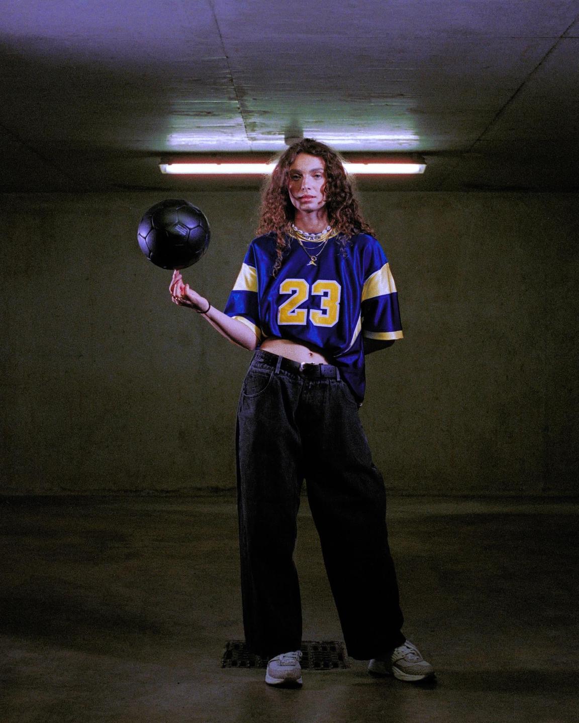 A woman in a blue and yellow jersey balances a black soccer ball on her finger in a dimly lit room.