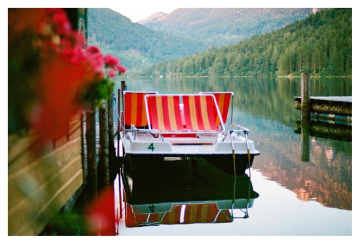 A boat with red and yellow striped seats is docked on a calm lake surrounded by mountains and trees.