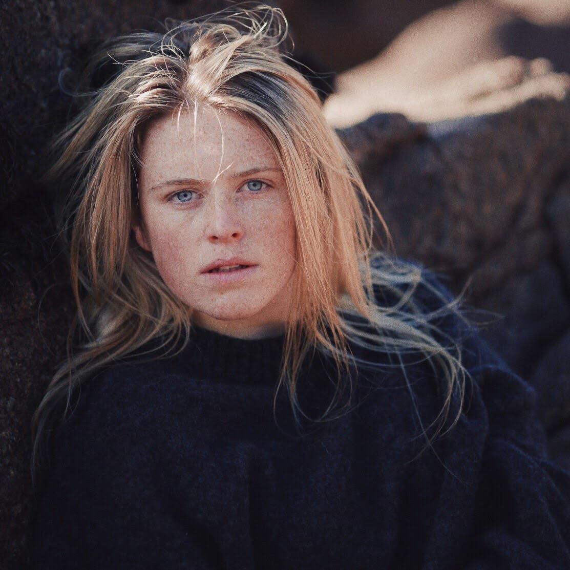 A woman with long blonde hair and freckles looks directly at the viewer.