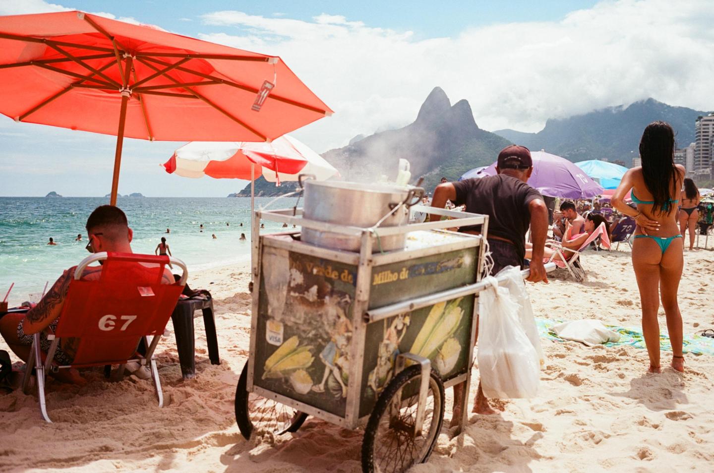 A man sells steamed corn from a cart on a crowded beach with mountains in the background.