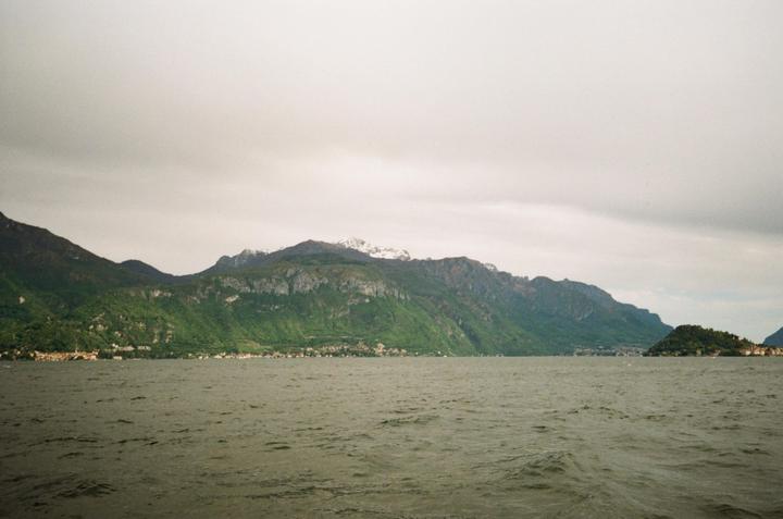 A wide body of water with small waves is in the foreground, with a long, green mountain range and a small town along the shore in the background under a cloudy sky.