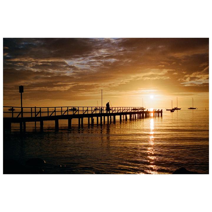 A person stands on a pier over water with boats as the sun sets, casting a golden glow.