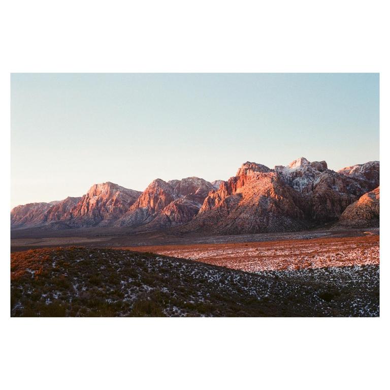 A mountain range with snow-dusted peaks is illuminated by golden light at sunset, with a vast, snow-covered plain in the foreground.